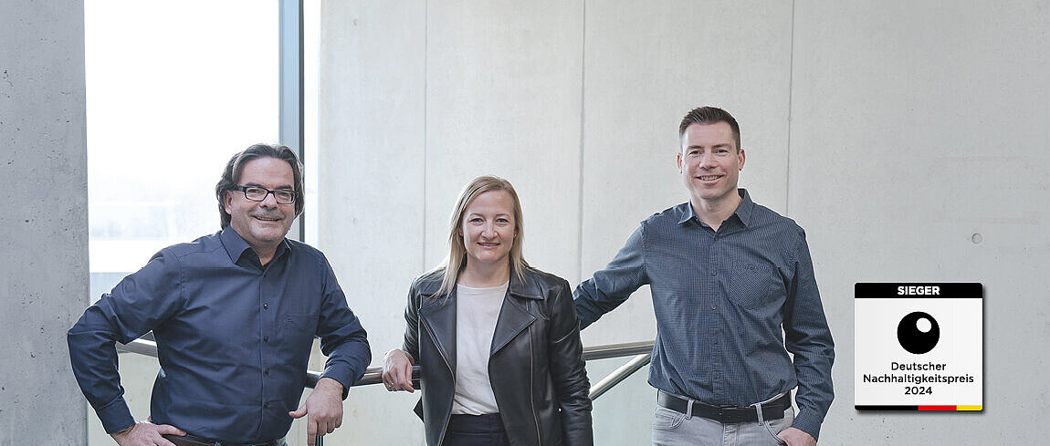 Daniel Büchle, Yvonne Cvilak und Mike Reif lehnen an einem Geländer und lächeln in die Kamera. Unten rechts befindet sich das Siegersiegel für den Deutschen Nachhaltigkeitspreis 2024.