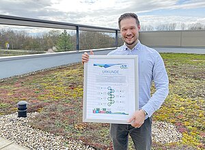 Sustainability Manager Christoph Teusch steht mit einer Urkunde auf einer Dachterrasse. Er trägt ein hellblaues Hemd und lächelt in die Kamera.