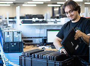 Ein Mann in schwarzem T-Shirt packt einen Laptop in eine Box auf einem Rollband. 