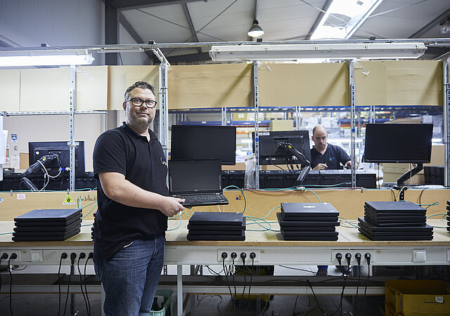 Ein Mann steht vor einer Werkbank, auf der sich viele Laptops stapeln. Er hält einen aufgeklappten Laptop in der Hand.