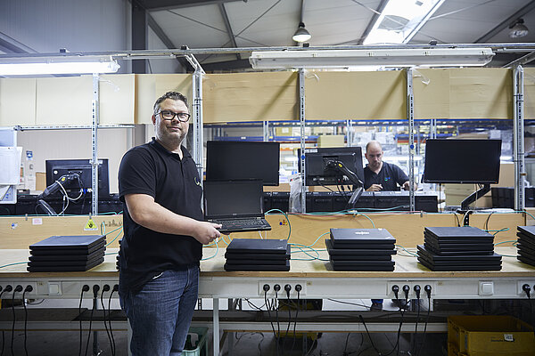 Ein Mann steht vor einer Werkbank, auf der sich viele Laptops stapeln. Er hält einen aufgeklappten Laptop in der Hand.