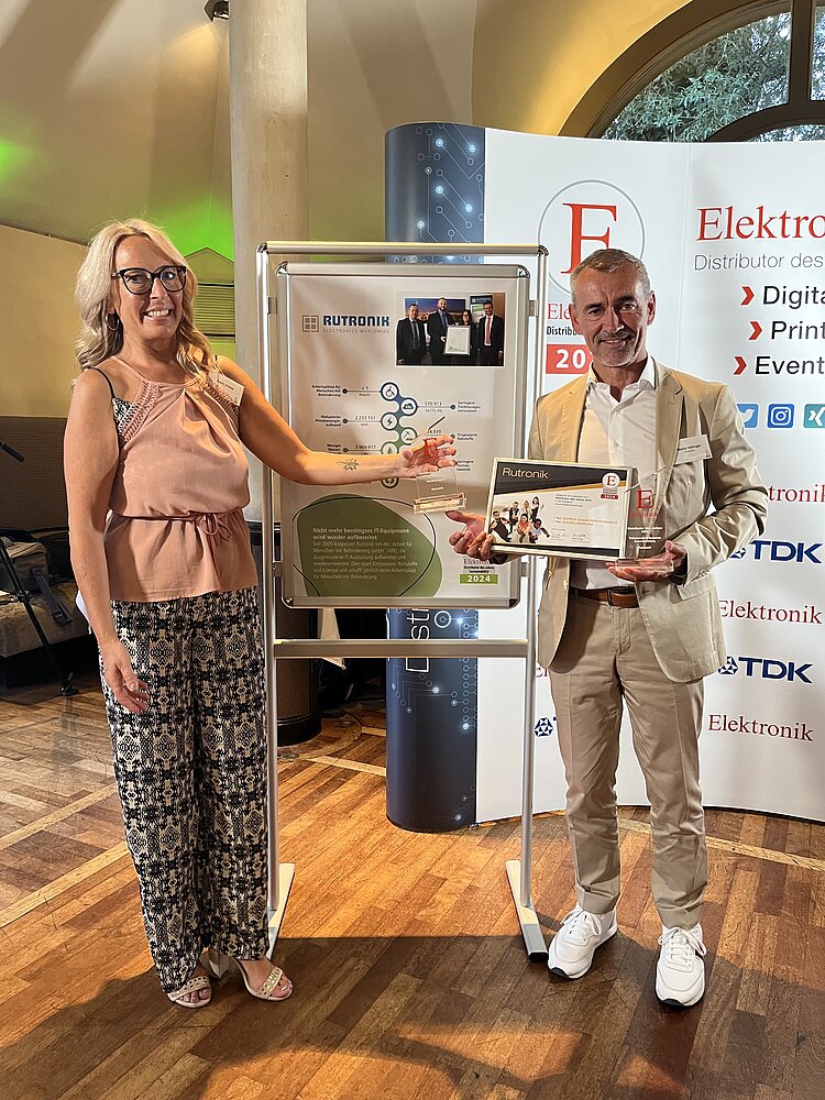 Eine blonde Frau in legerer Kleidung überreicht Marco Nabinger von Rutronik den Sonderpreis Nachhaltigkeit. Herr Nabinger trägt einen weißen Anzug. Die beiden stehen vor einer Wand mit Logos und einem Poster, auf dem grafische Elemente aus der AfB-Wirkungsurkunde kombiniert worden sind.