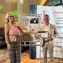 Eine blonde Frau in legerer Kleidung überreicht Marco Nabinger von Rutronik den Sonderpreis Nachhaltigkeit. Herr Nabinger trägt einen weißen Anzug. Die beiden stehen vor einer Wand mit Logos und einem Poster, auf dem grafische Elemente aus der AfB-Wirkungsurkunde kombiniert worden sind.