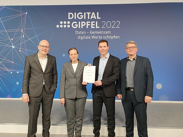 4 Personen in Anzügen stehen vor einer blauen Wand mit der Aufschrift "Digitalgipfel 2022. Daten - Gemeinsam digitale Wrte schöpfen".