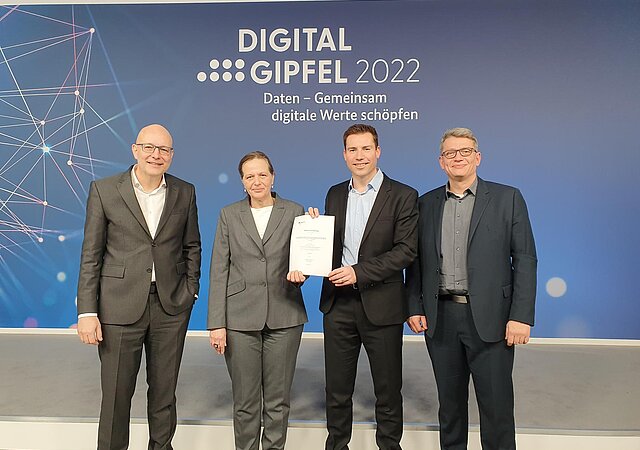 4 Personen in Anzügen stehen vor einer blauen Wand mit der Aufschrift "Digitalgipfel 2022. Daten - Gemeinsam digitale Wrte schöpfen".