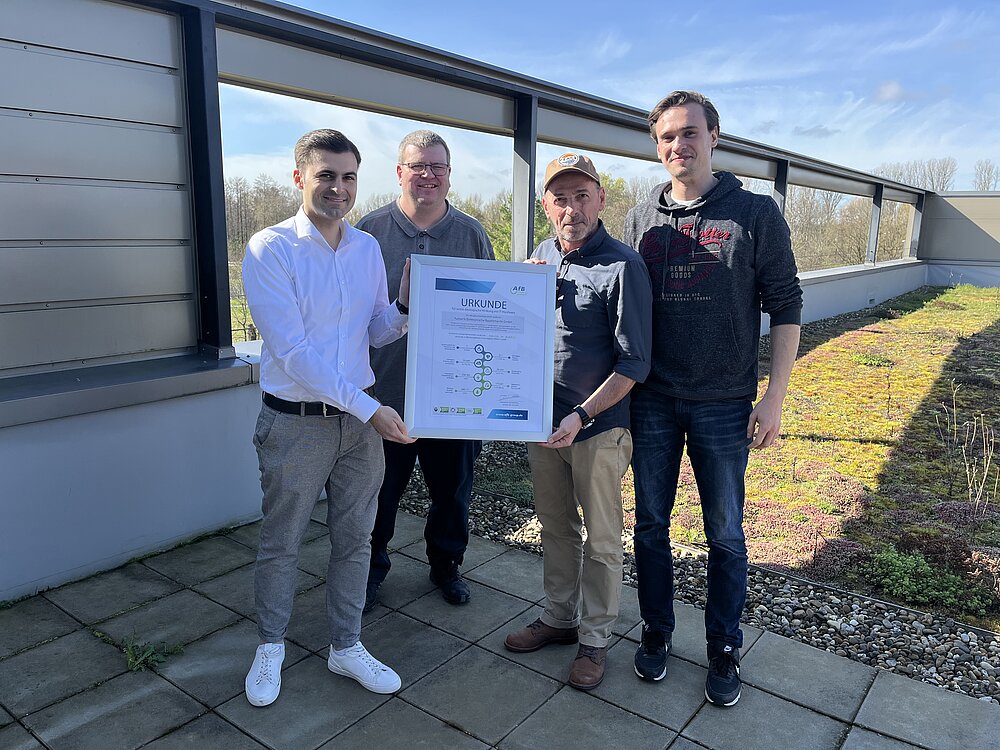 Vier Männer stehen auf der Dachterrasse bei AfB. Ganz links steht Oliver Raguse, Partnermanager bei AfB und überreicht drei Herren von Rutronik die Wirkungsurkunde für das Jahr 2023. Es ist ein frühlingshafter Wetter bei blauem Himmel, die Männer tragen legere Kleidung.